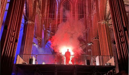 Eine dunkle Gestalt steht auf einem Gerüst im Nebel in einem Kirchenschiff.