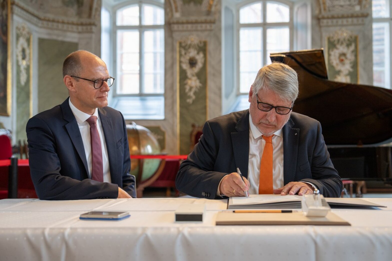 Zwei Männer sitzen hinter einem Tisch mit weißer Tischdecke. Der rechte Mann unterzeichnet ein Dokument.