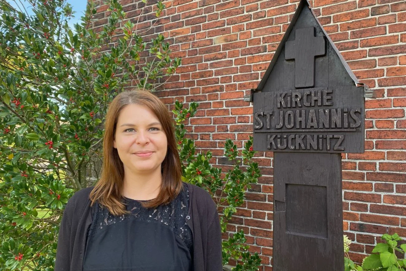 Junge Frau steht vor einem Busch, neben ihr ist ein hölzernes Kreuz