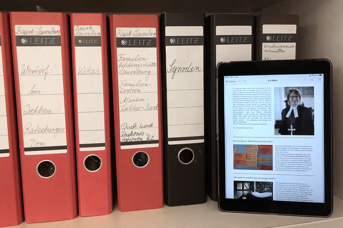 Ein iPad steht vor mehreren schwarzen und roten Aktenordner und zeigt Ausschnitte aus dem Newsletter "Kirchenkreis informiert"