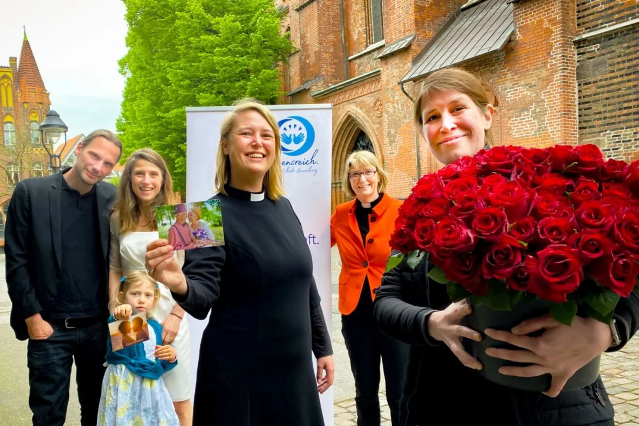 Sechs Menschen schauen freundlich in die Kamera: Links hinten im Bild ein Brautpaar mit einem Blumenkind. Hinten rechts eine Pastorin neben einem Aufsteller. Im Vordergrund halten zwei Pastorinnen einen großen Strauß halbaufgeblühter roter Rosen und Segenspostkarten in die Kamera.
