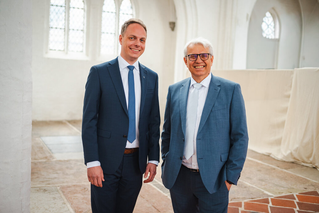 Zwei Männer stehen nebeneinander: Der linke mit weißem Hemd, hellblauer Krawatte und dunkelblauem Anzug, der rechte trägt ein weißes Hemd und ein hellblaues Sako. Im Hintergrund eine leere Kirche mit weißen Wänden und Boden aus Ziegelsteinen.