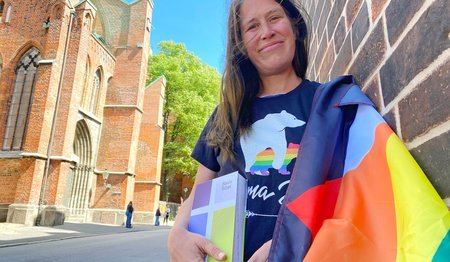 Eine Frau mit einer Bibel und einer Regenbogenflagge lehnt an einer Hauswand. 