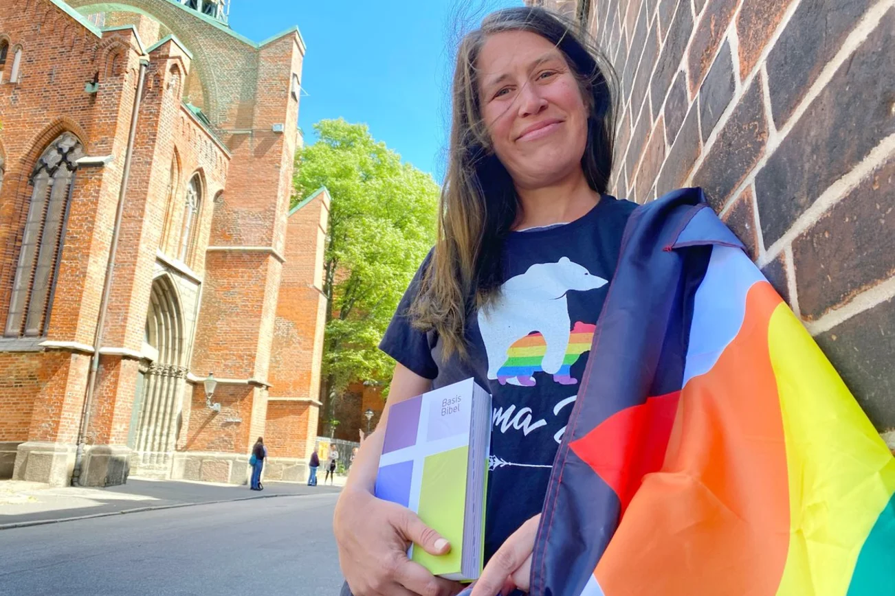 Eine Frau mit einer Bibel und einer Regenbogenflagge lehnt an einer Hauswand. 