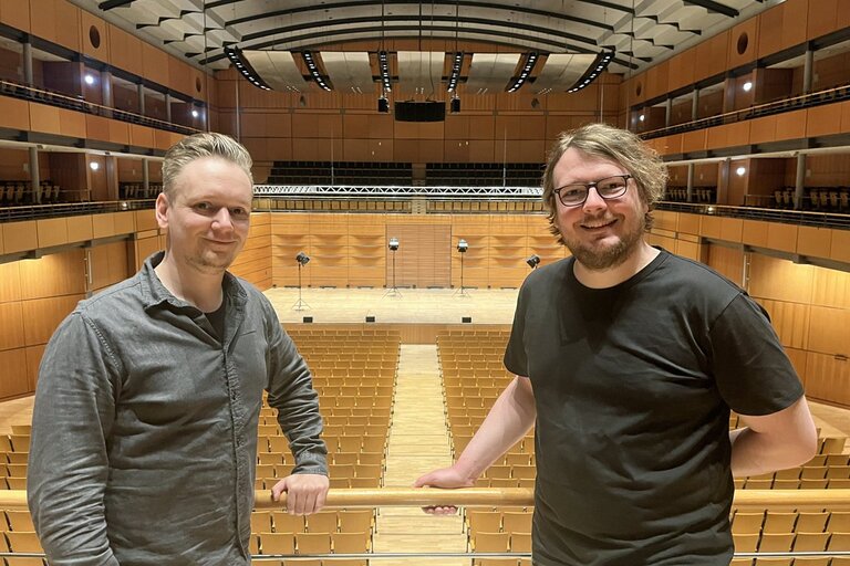 Zwei Männer stehen auf in einem leeren Konzertsaal.