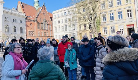 Eine Gruppe Menschen steht auf dem Koberg bei blauen Himmel, im Hintergrund die Häuserfassaden Lübecks 