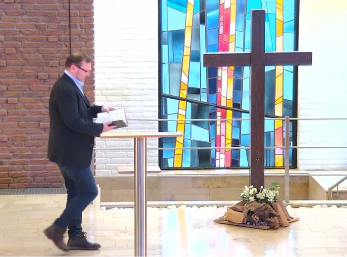 Ein Mann geht zu einem Stehpult, er hält ein offenes Buch in den Händen, rechts steht ein großes Holzkreuz, im Hintergrund ist ein buntes Kirchenfenster zu sehen