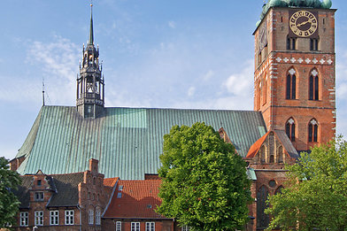 St.-Jakobi-Kirche Lübeck vom Klingenberg fotografiert. Der Turm ragt in den blauen Himmel. Es ist eine Seitenansicht der Kirche, rechts der Turm, links das Kirchenschiff. Vor der Kirche stehen links zwei Bäume mit grünen Blättern. - Copyright: Ev.-Luth. Kirchenkreis Lübeck-Lauenburg