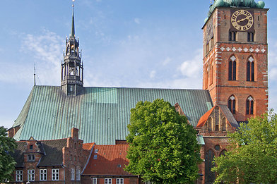 St.-Jakobi-Kirche Lübeck vom Klingenberg fotografiert. Der Turm ragt in den blauen Himmel. Es ist eine Seitenansicht der Kirche, rechts der Turm, links das Kirchenschiff. Vor der Kirche stehen links zwei Bäume mit grünen Blättern. - Copyright: Ev.-Luth. Kirchenkreis Lübeck-Lauenburg