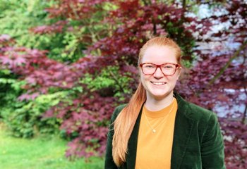 Eine Frau mit langen Haaren, Brille, grüner Jaclke und orangenem Oberteil schaut in die Kamera. Im Hintergrund eine Hecke mit grünen und roten Blättern - Copyright: Foto: privat