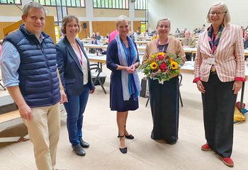 Fünf Personen stehen im Halbkreis. Sie blicken freundlich in die Kamera. Eine Frau hält einen Blumenstrauß in der Hand. Im Hintergrund ein eine Sporthalle mit Menschen, sitzend an Tischen. - Copyright: Bastian Modrow