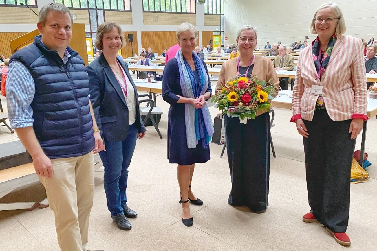Fünf Personen stehen im Halbkreis. Sie blicken freundlich in die Kamera. Eine Frau hält einen Blumenstrauß in der Hand. Im Hintergrund ein eine Sporthalle mit Menschen, sitzend an Tischen.