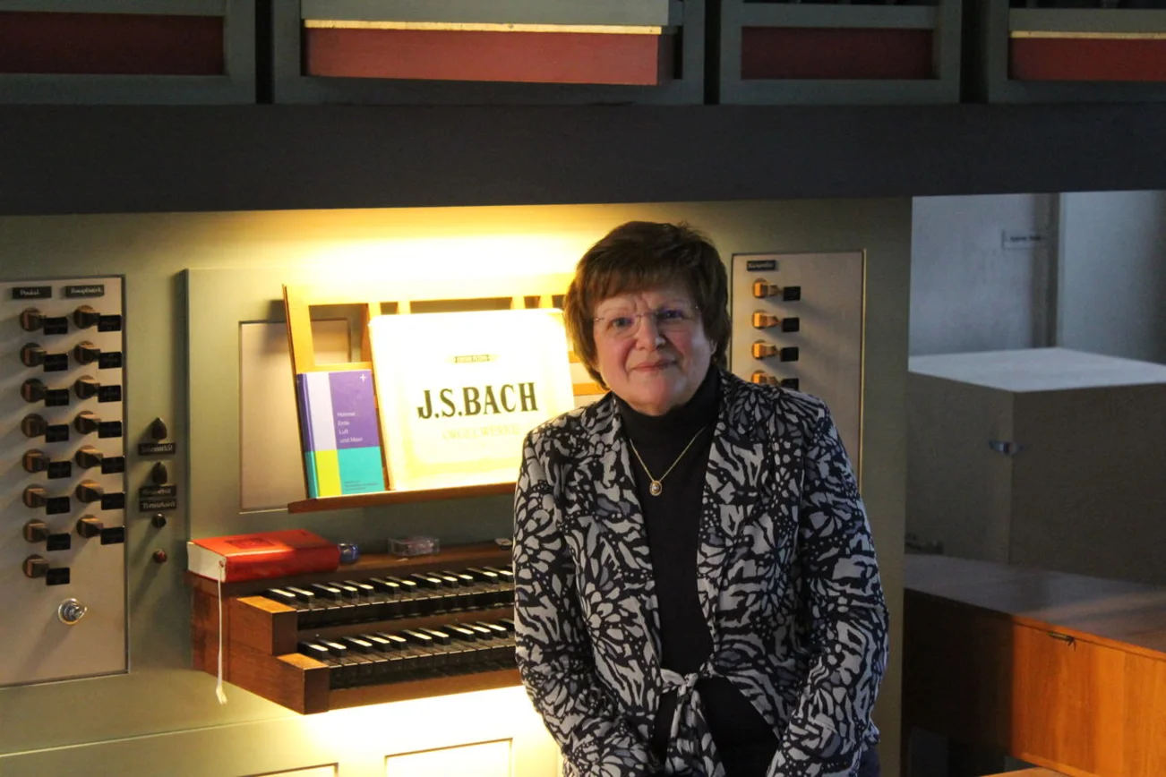 Eine Frau mit dunklen, kurzen Haaren sitzt an einem Orgelspieltisch und blickt freundlich in die Kamera. Auf dem Notenpult liegt ein Notenheft von Johann Sebastian Bach.