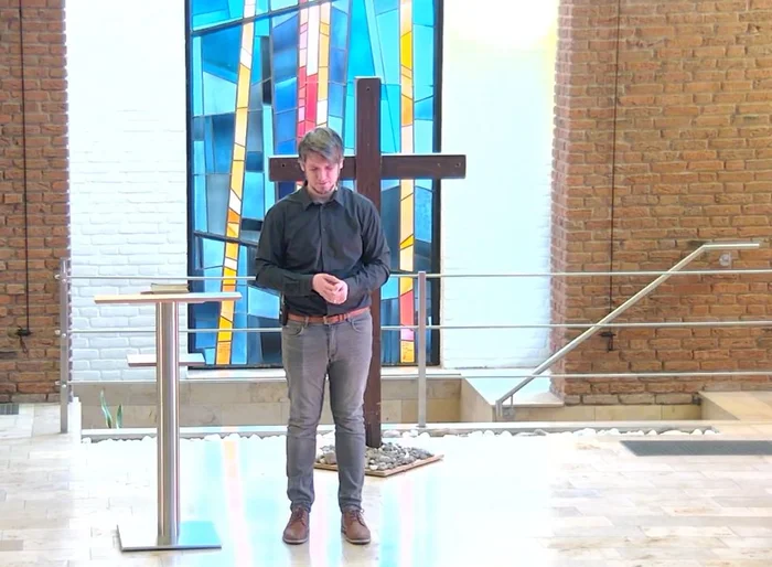 Ein Mann steht betend vor einem Holzkreuz und hat die Augen geschlossen - Neben ihm steht ein Stehtisch mit einer Bibel darauf. hinter ihm ist ein Becken und ein Handlauf zu sehen, ebenso ein buntes Glasfenster