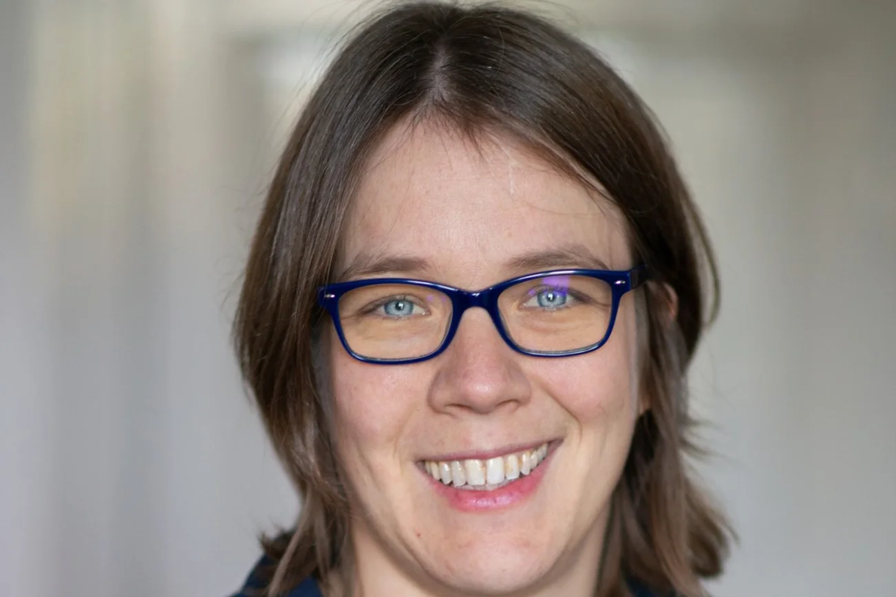 Das Portait einer Frau mit Brille und kinnlangen Haaren. Sie lächelt freundlich .