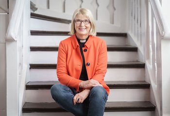 Eine Frau in Jeans, orangem Blazer und Collarhemd sitzt auf den Stufen einer weißen Holztreppe. Sie blickt freundlich in die Kamera. - Copyright: Guido Kollmeier