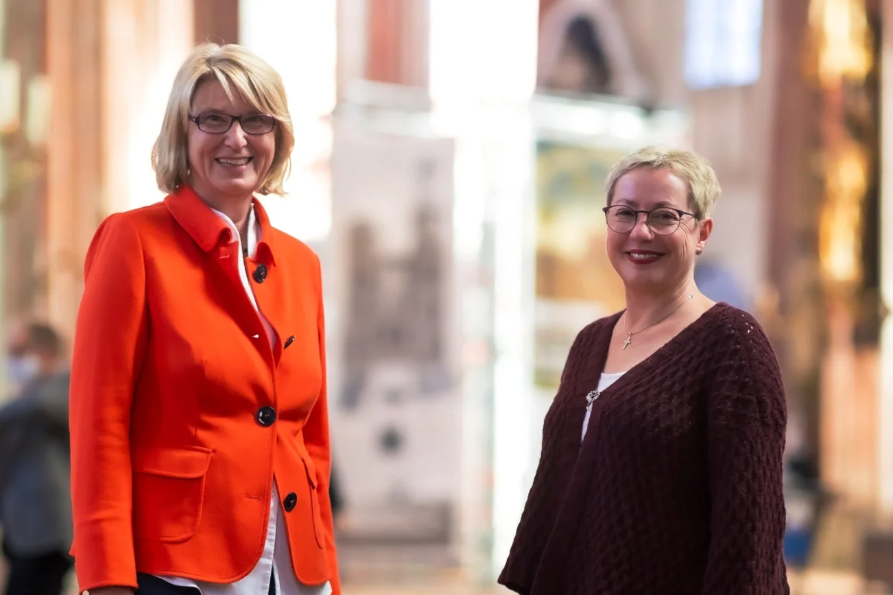 Zwei Frauen im Halbportrait schauen freundlich in die Kamera. Sie sind einander zugewandt, im Hintergrund ist unscharf der Kirchraum von St. Marien zu erkennen.