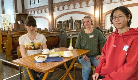 Drei Frauen sitzen in einer Kirche an einem Tisch. 