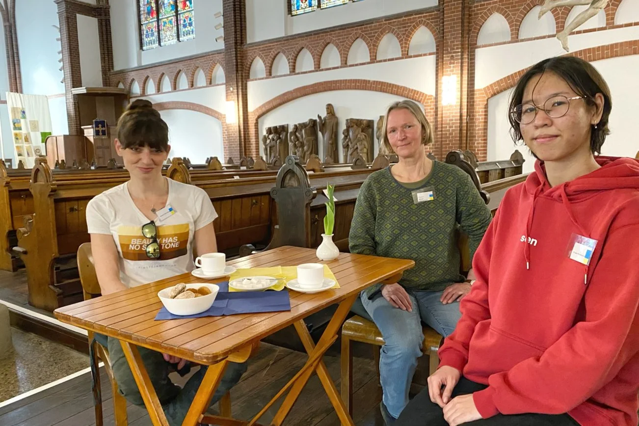 Drei Frauen sitzen in einer Kirche an einem Tisch. 