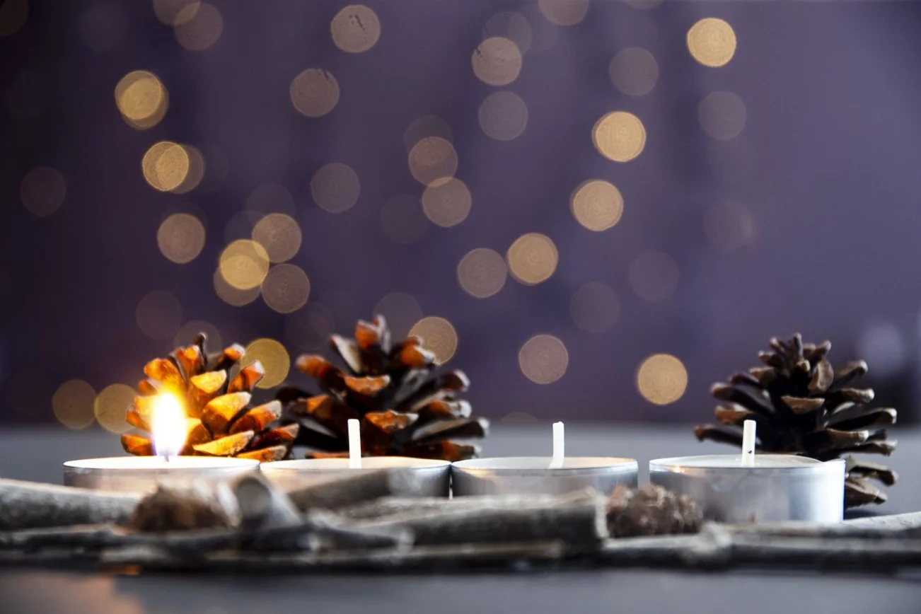 Vier Teelichter stehen im Vordergrund und eines leuchtet. Im Hintergrund sind Tannenzapfen und undeutliche Lichter