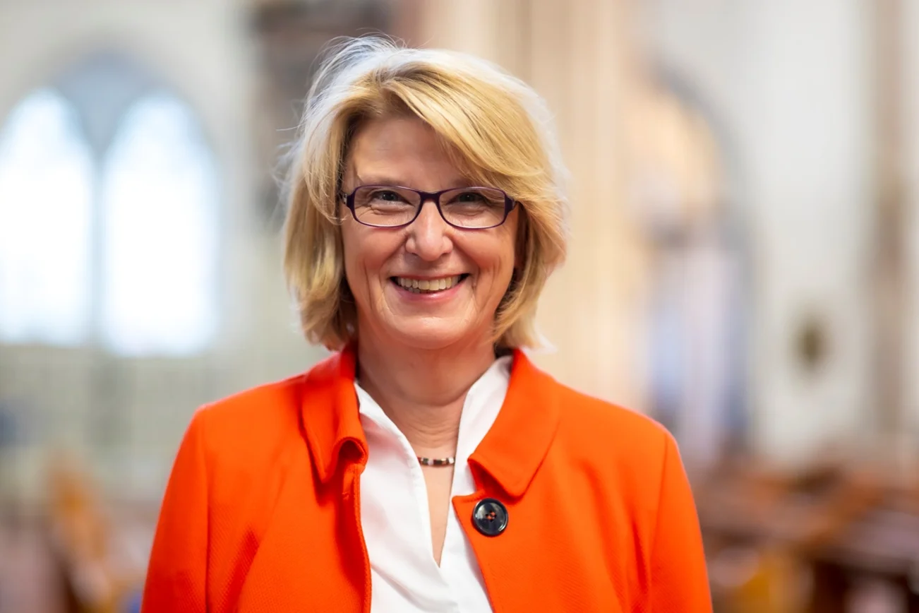 Eine Frau im Portrait. Sie trägt eine weiße Bluse, darüber eine Jacke in orange.