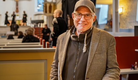 Ein Mann mit Brille, Sakko und Basecap steht in einer Kirche und lächelt in die Kamera.