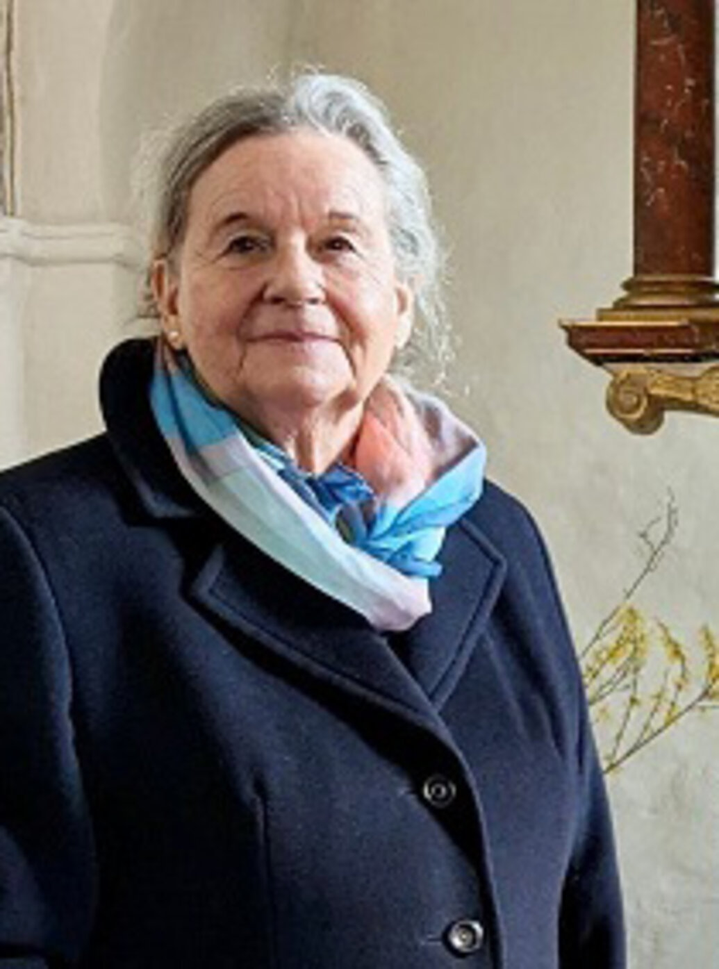 Eine Dame mit grauen Haaren trägt einen dunkelblauen Mantel und ein pastellfarbenes Tuch um den Hals