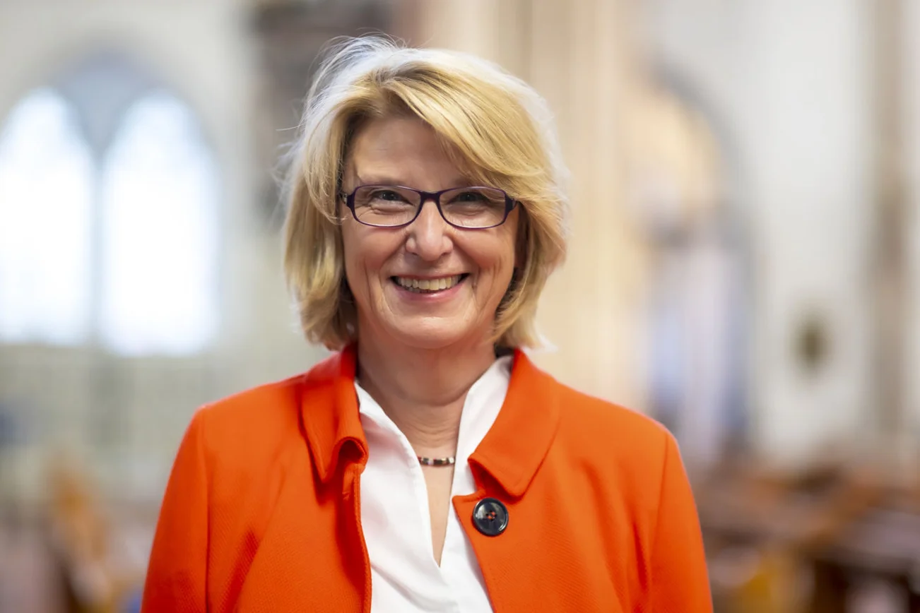 Eine blonde Frau mit Brille und orangefarbener Jacke lächelt freundlich in die Kamera