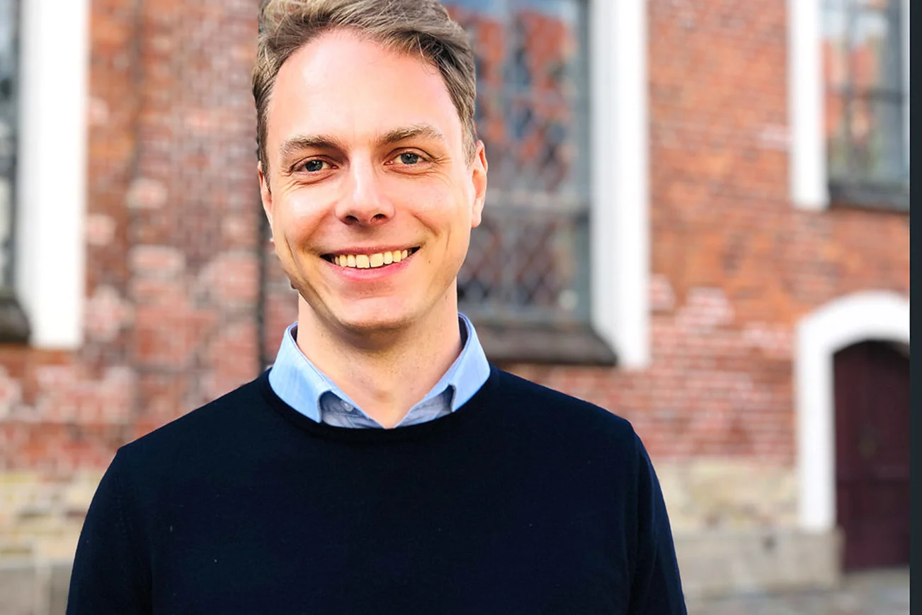 Junger Mann steht lächelnd vor einer Backsteinkirche.