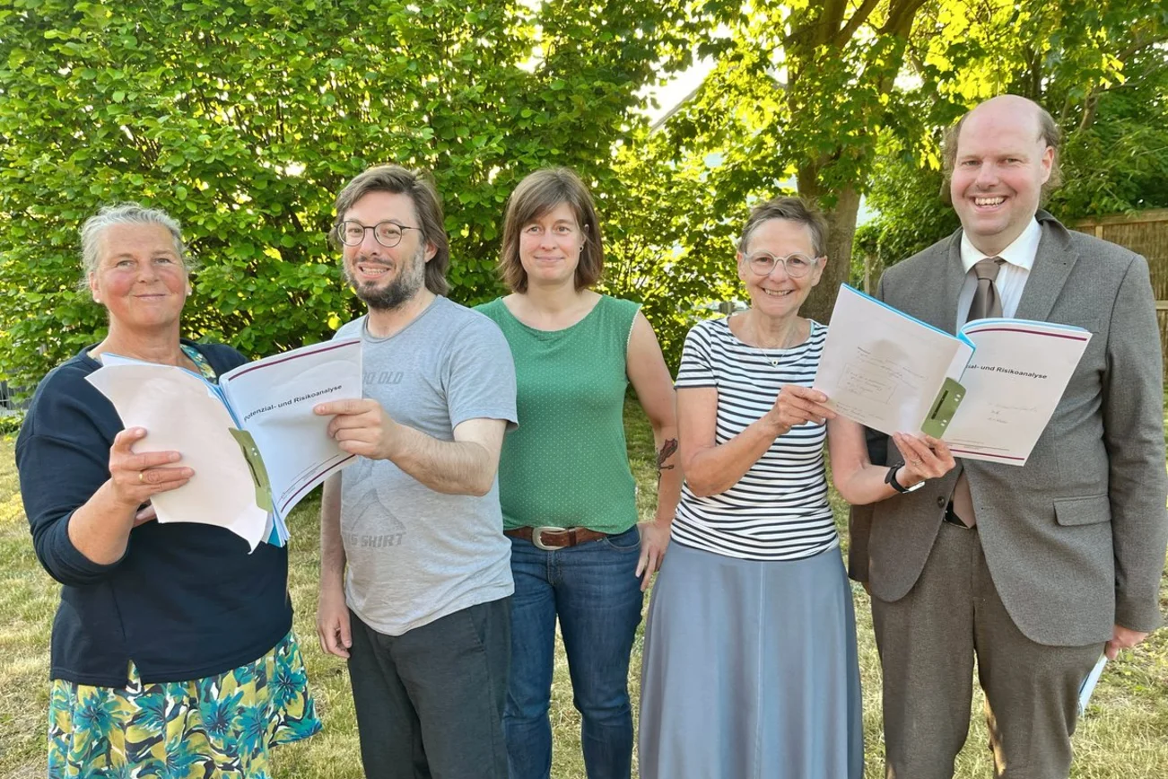 Menschen stehen in einem Garten und blicken in Aktenordner. 