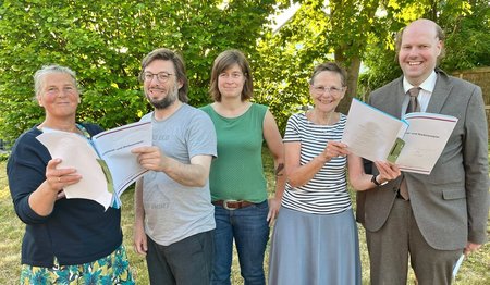 Menschen stehen in einem Garten und blicken in Aktenordner. 