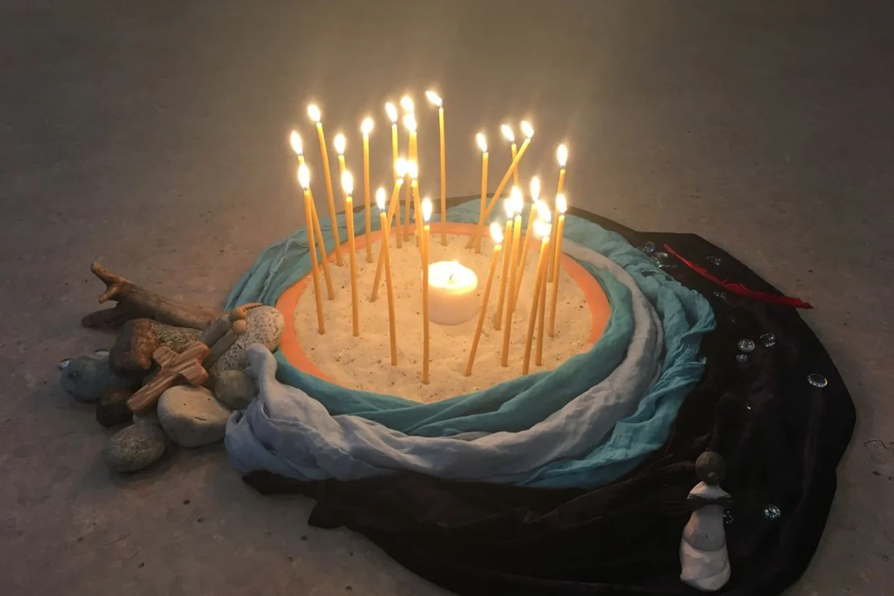 Auf einem sandigen Untergrund ist ein Kreis aus Tüchern gelegt. Darin stehen brennende Kerzen. An der Seite liegen Steine, ein Holzkreuz und Figuren.