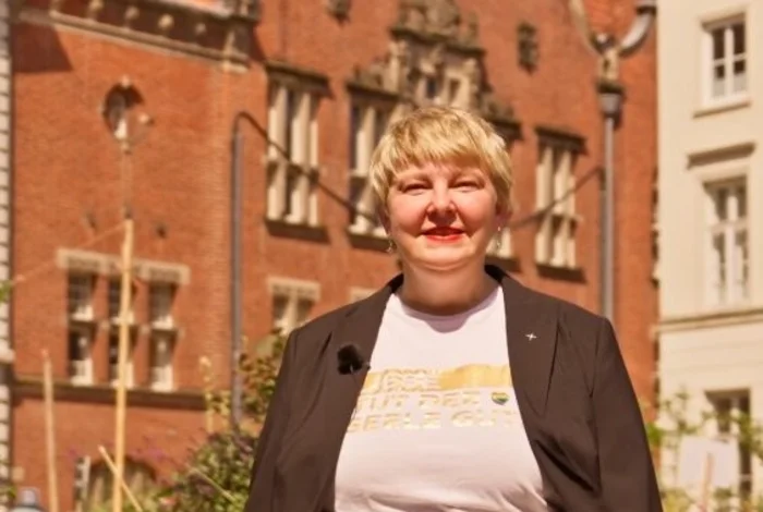 Eine Frau mit kurzen blonden Haaren steht zwischen Pflanzen in einer Altstadt.