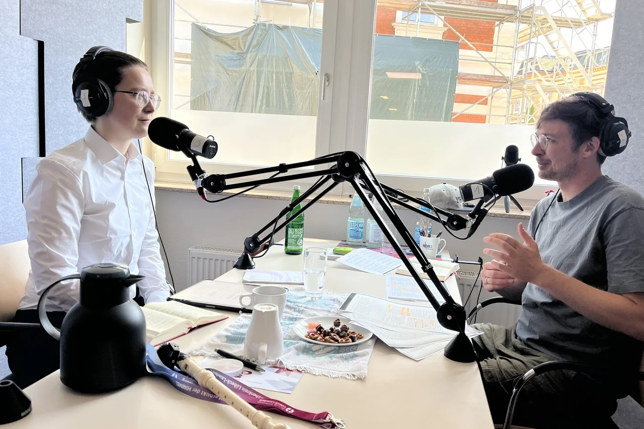 Zwei Männer sitzen in einem Podcaststudio an Mikrofonen und reden.