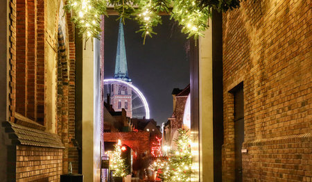  Weihnachtlich geschmückter Gang am Weihnachtsmarkt St. Jakobi. Tannen mit hellen Lichterketten ragen von oben herab. Im Hintergrund ist das fahrende Riesenrad zu sehen, dahinter Der beleuchtete Turm von St. Jakobi.