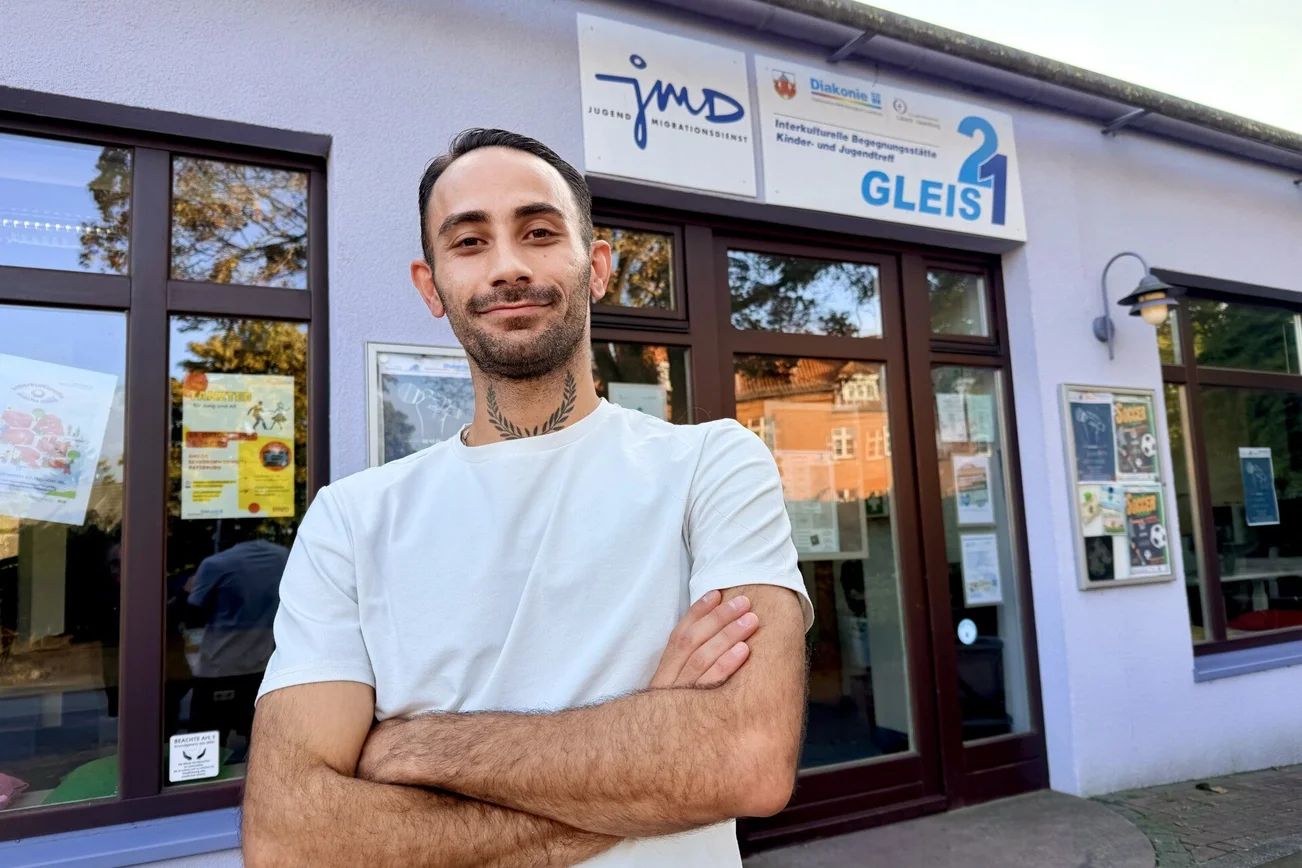 Ein Mann mit weißem T-Shirt und dunklen kurzen Haaren und Drei-Tage-Bart steht vor einem Gebäude mit großen Türen und Fenstern. Er hat die Arme vor sich verschränkt.