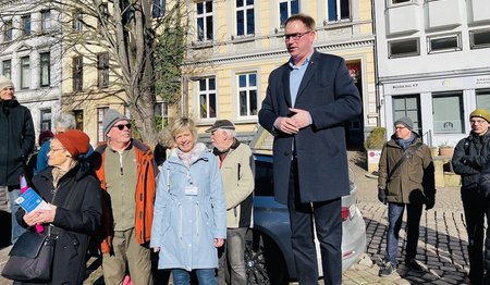 Lübecks Bürgermeister Jan Lindenau steht erhöht auf einem Stein, eine Gruppe Menschen steht links und rechts von ihm. Ein silbernes Auto steht hinter ihm. Im Hintergrund ist eine gelbe und graue Häuserfront zu sehen. Ein Baum ohne Blätter steht hinter der linken Menschengruppe.
