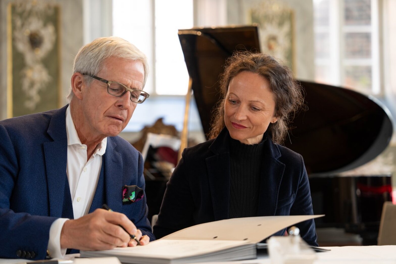 Ein Mann, links im Bild unterzeichnet ein Dokument, die Frau rechts von ihm schaut dabei zu.