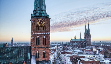 Die Dächer von Lübeck sind leicht von Schnee bedeckt. Auf der linken Seite sieht man den Turm von St. Jakobi, leicht versetzt nach rechts hinten, ist St. Marien zu sehen und hinter St. Marien, sieht man die Turmspitze von St. Petri. Im Hintergrund links sieht man klein den Turm von St. Aegidien.