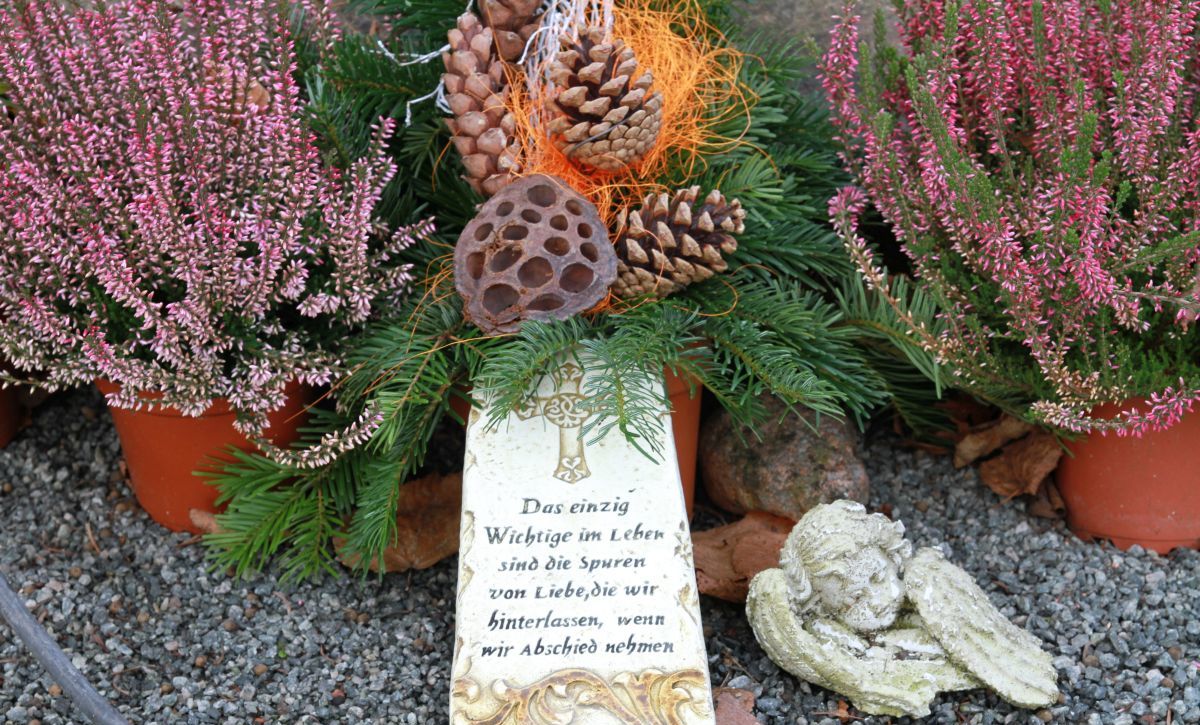 Ein Grab mit Heideblumen, einem beschriebenen hellen Stein und einem kleinen Engel davor. 
