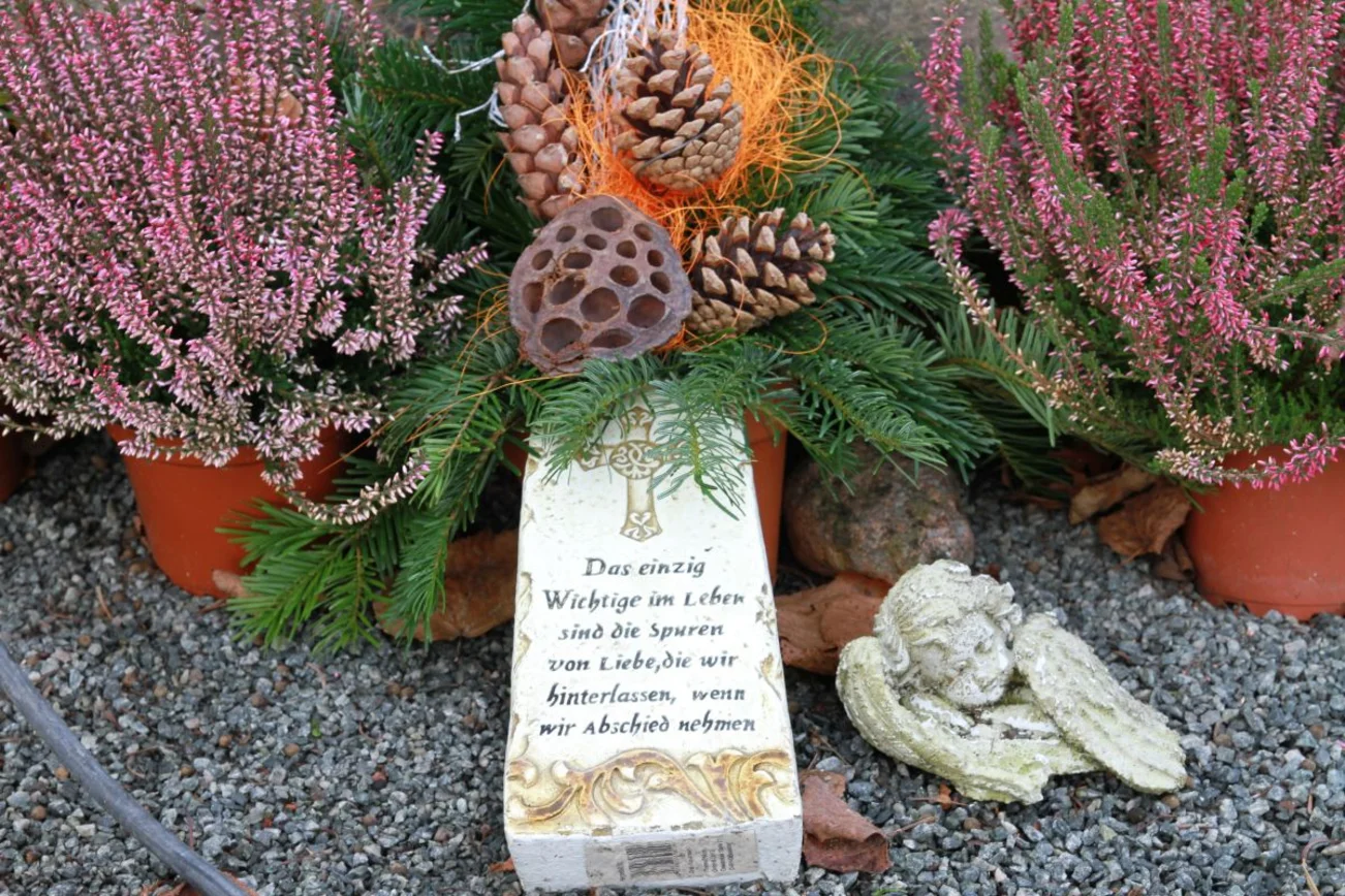 Ein Grab mit Heideblumen, einem beschriebenen hellen Stein und einem kleinen Engel davor. 