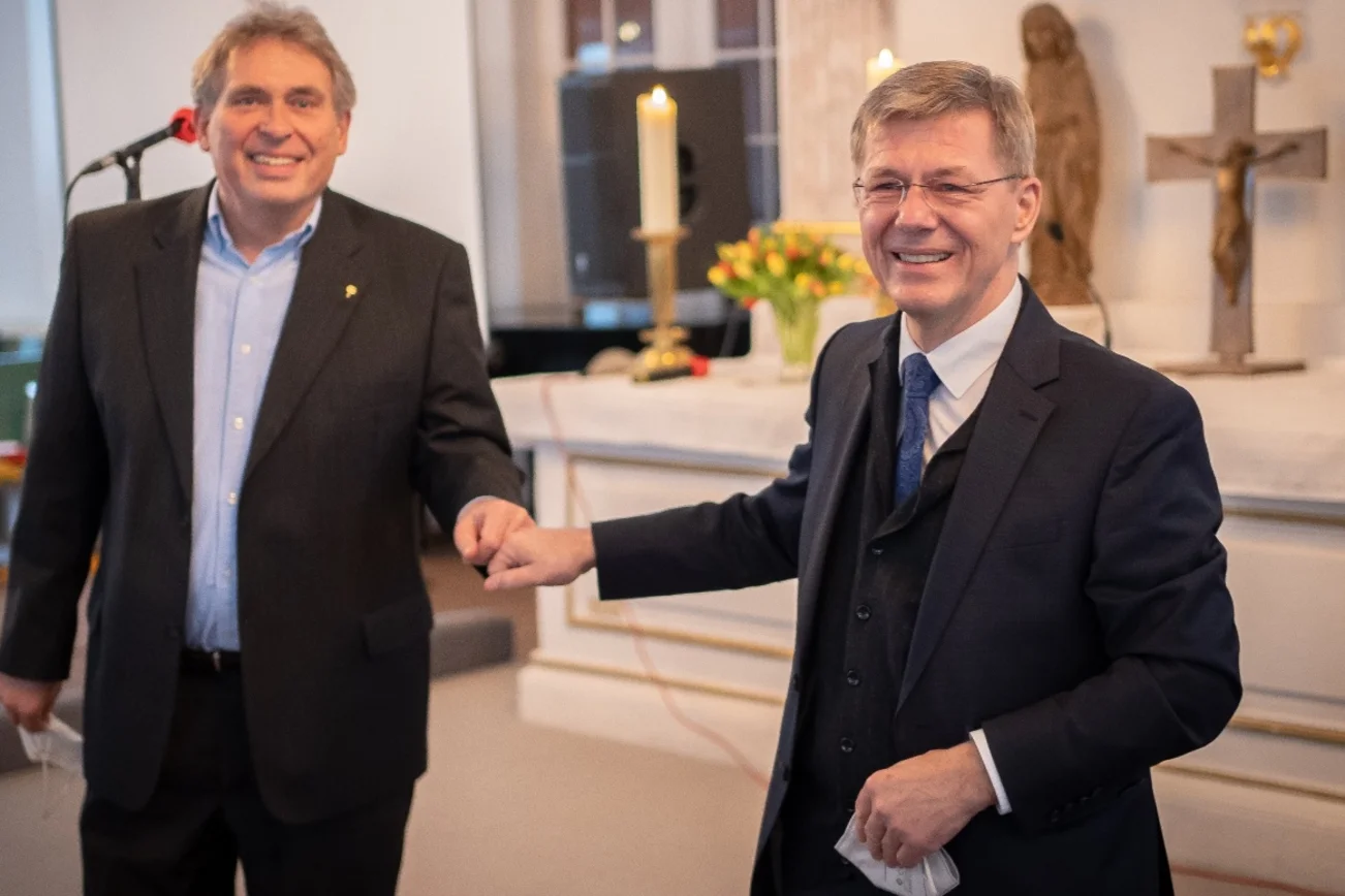 Zwei Männer stehen auf Abstand in einem Kirchraum und geben sich die Corona-Faust.