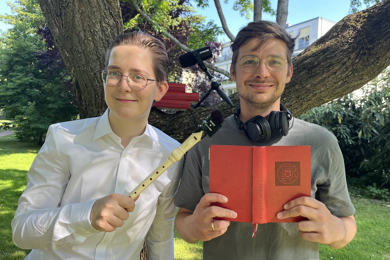 Zwei Männer stehen vor einem Baum und halten eine Blockflöte und ein rotes Gesangbuch.