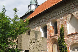 Ein Steinkreuz mit einer Abbildung der Sonne, davor eine Sichel mit einem Büschel Getreide.