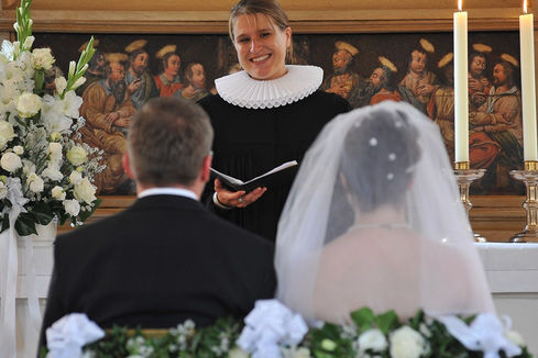 Hochzeitspaar sitzt auf geschmückten Stühlen mit Blick auf die Pastorin, diese hält ein offenes Buch in der Hand, steht vor dem Altar und lächelt das Brautpaar an  - Copyright: Ev.-Luth. Kirchenkreis Lübeck-Lauenburg