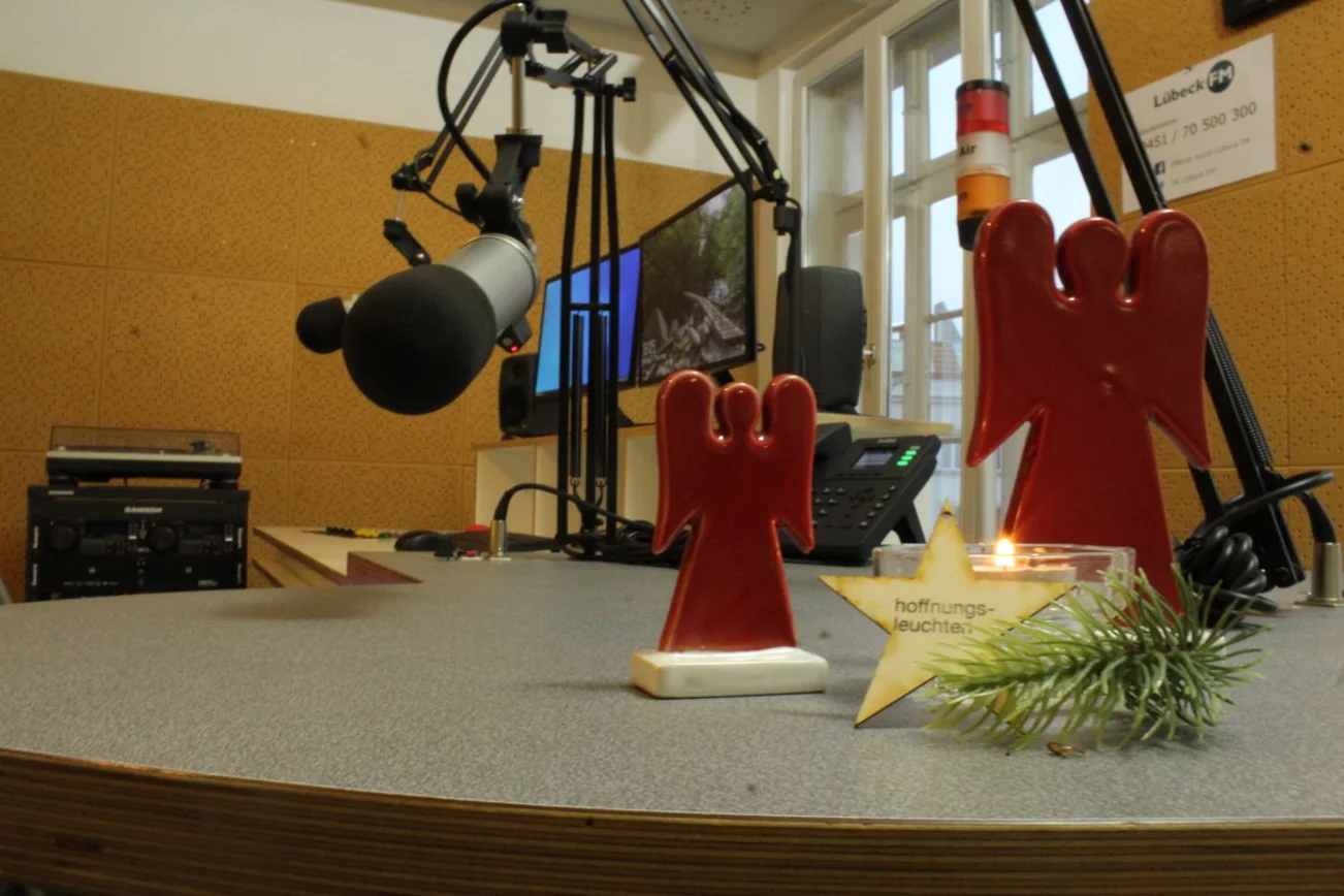 Im Vordergrund sieht man zwei stilisierte rote Engel hinter einer brennenden Kerze mit einem Tannenzweig und einem Stern. Im Hintergrund sieht man ein Radiostudio.