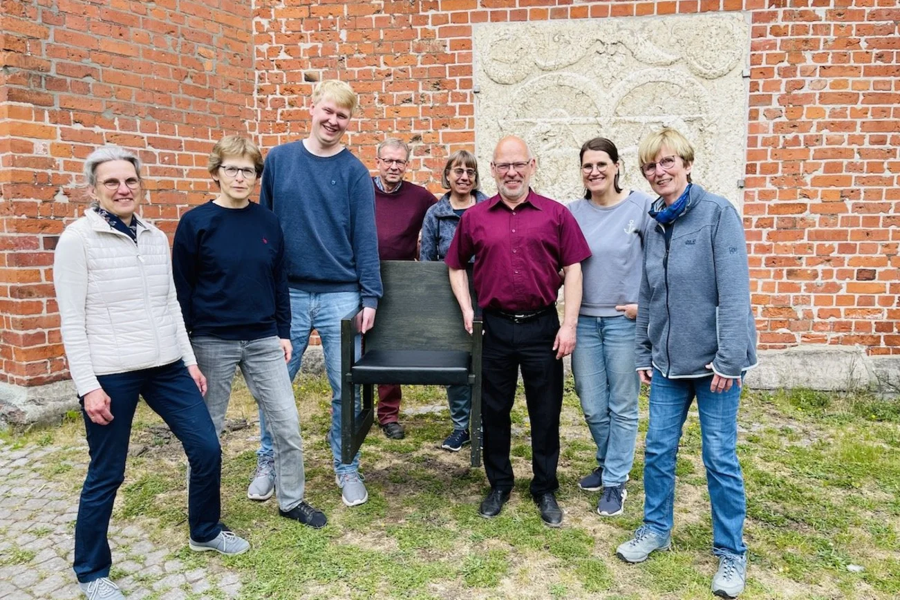 Das Organisations-Team des Dom-Tages lädt alle kleinen und großen Menschen, kirchlich oder nicht, zum Dom-Tag am 21. Juni ein: Karin Harder, Isabel Kriegeskotten, Per Olsen, Thomas Hornecker, Silke Luhmann, Martin Klatt, Margrit Wegner und Cornelia Schäfer (v.li.). Sie stehen vor dem Dom, im Hintergrund sind die dicken Kirchenmauern zu sehen, und halten symbolische einen Kirchenstuhl in den Händen.  