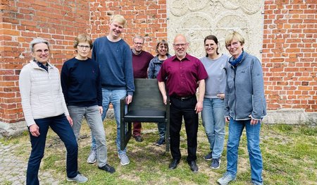 Das Organisations-Team des Dom-Tages lädt alle kleinen und großen Menschen, kirchlich oder nicht, zum Dom-Tag am 21. Juni ein: Karin Harder, Isabel Kriegeskotten, Per Olsen, Thomas Hornecker, Silke Luhmann, Martin Klatt, Margrit Wegner und Cornelia Schäfer (v.li.). Sie stehen vor dem Dom, im Hintergrund sind die dicken Kirchenmauern zu sehen, und halten symbolische einen Kirchenstuhl in den Händen.  