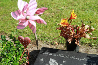 Grabstein mit der Aufschrift: "Ich bin ein Kind der Sterne, geheimnisvoll", neben dem eine rosa Windmühle und ein kleines rotes Herz mit weißen Punkten steckt. Eine Blumenvase mit Blumen steht am Kopfende des Grabsteins.