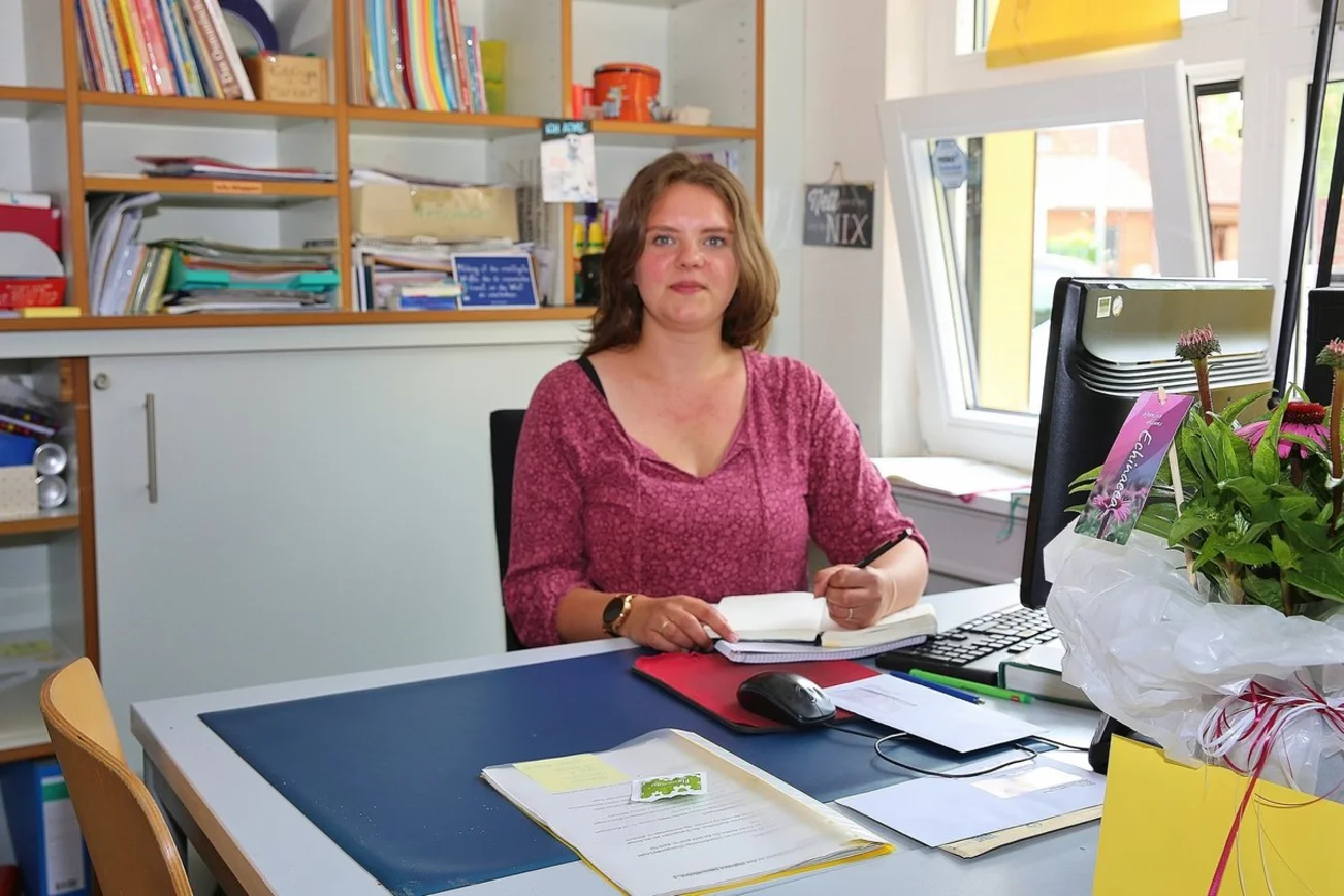 Eine junge Frau mit langen Haaren sitzt an einem Schreibtisch und schaut in die Kamera.